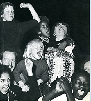 Archival black and white photograph of a lively group involving adults and children in a drama activity.