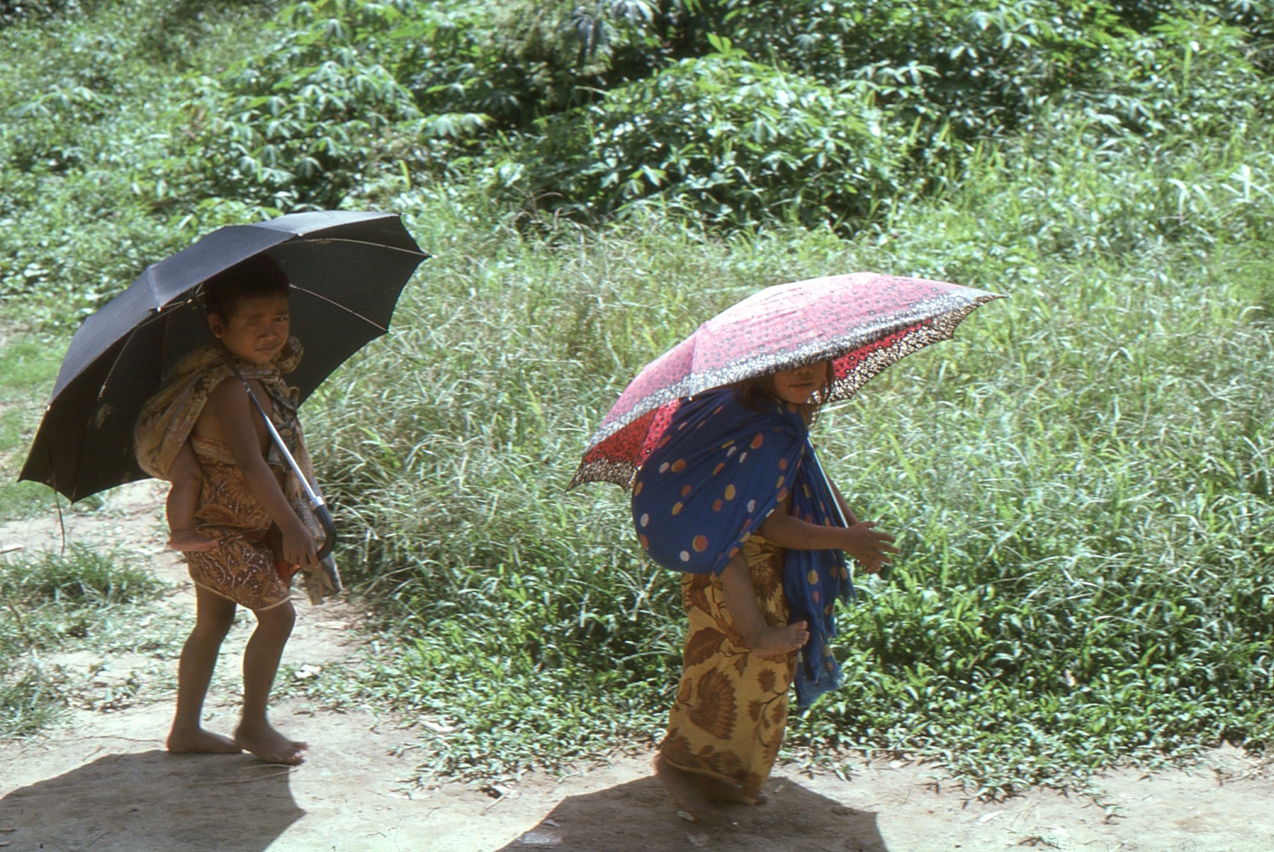 Two Temiar children caring for younger siblings while holding borrowed umbrellas in Kelantan in the 1970s